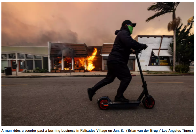 A man rides a scooter past a burning business in Palisades Village on Jan. 8. (Brian van der Brug / Los Angeles Times)